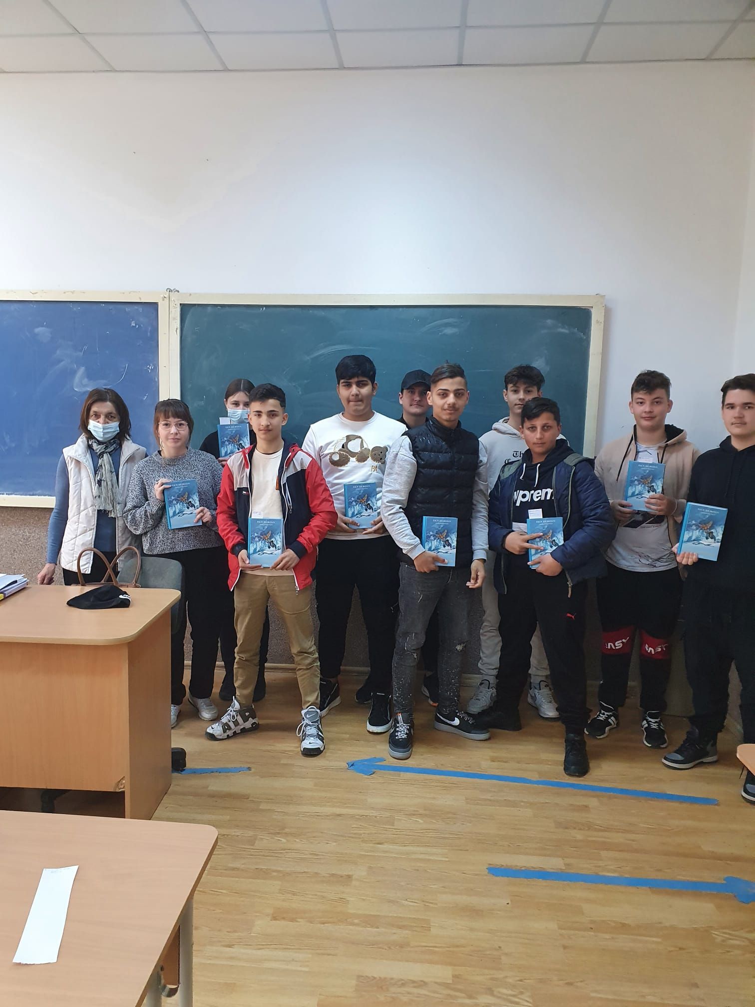 Grup de elevi de gimnaziu cu cărți albastre în mâini, în fața tablei
