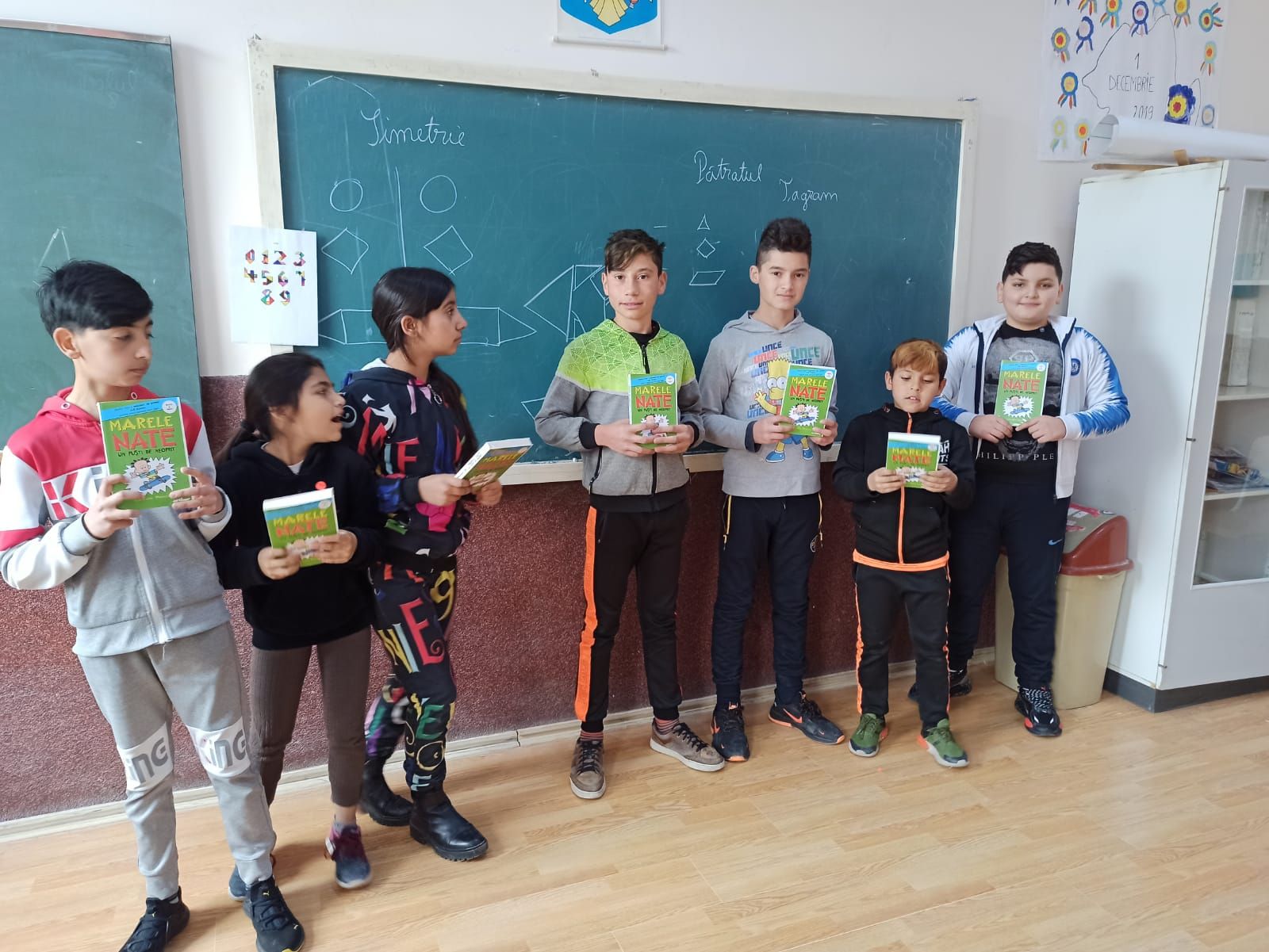 Elevi cu cărți în mâini, în fața tablei pe care sunt desenate figuri geometrice