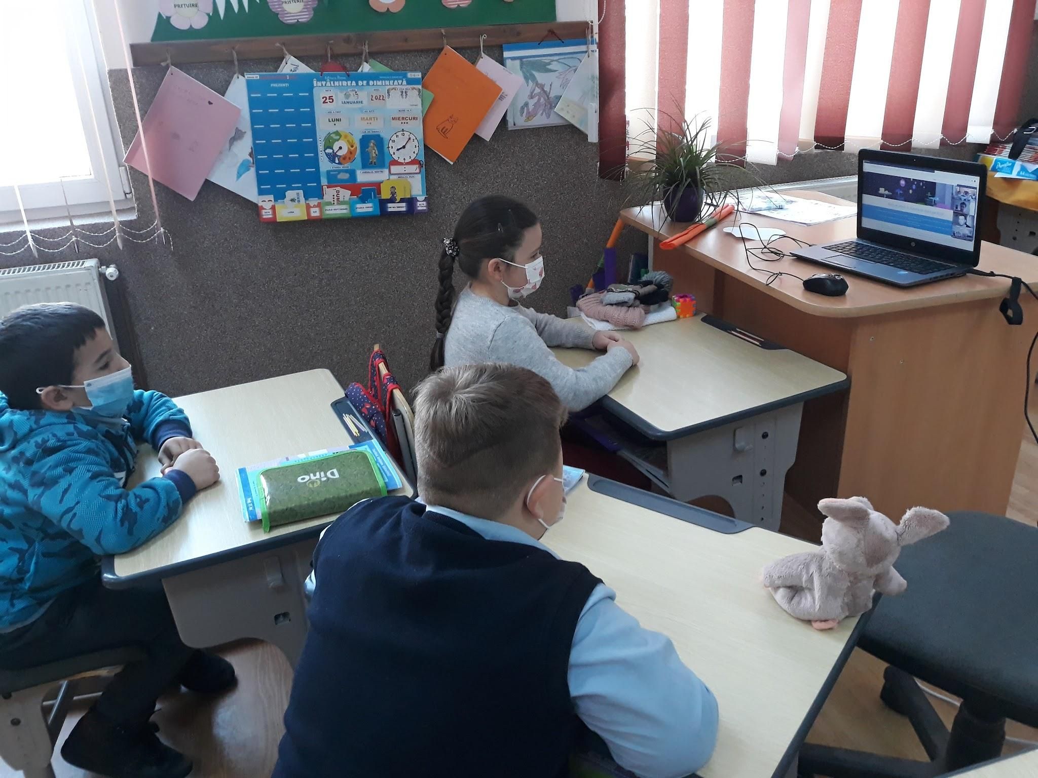 Elevi cu măști de protecție urmărind o lecție online pe laptopul de pe catedră
