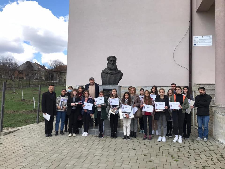 Participanții premiați, cu diplomele în mâini, în fața unei școli, lângă un bust