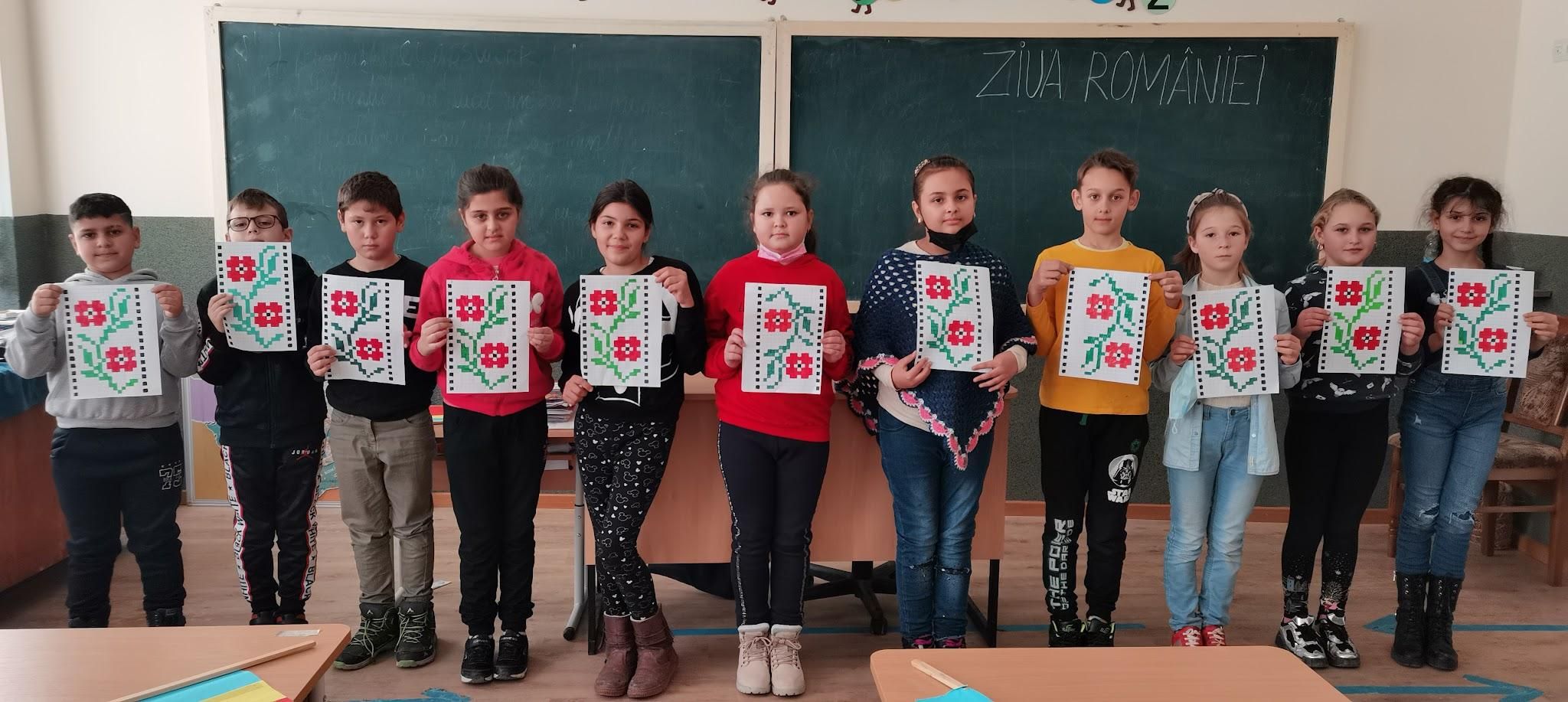 Elevi cu desene cu motive populare, în fața tablei pe care scrie „Ziua României”